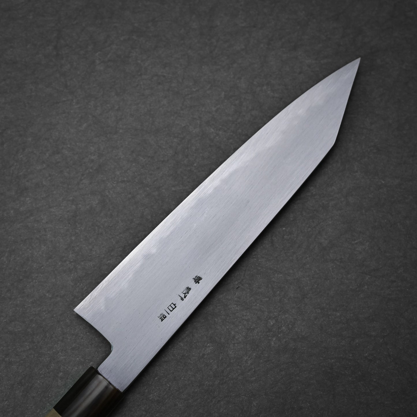 The Yoshikazu Tanaka shirogami#1 kiritsuke gyuto 225mm, featuring a pointed blade and Japanese characters near the handle, is crafted by Yoshikazu Tanaka and displayed on a dark textured surface.