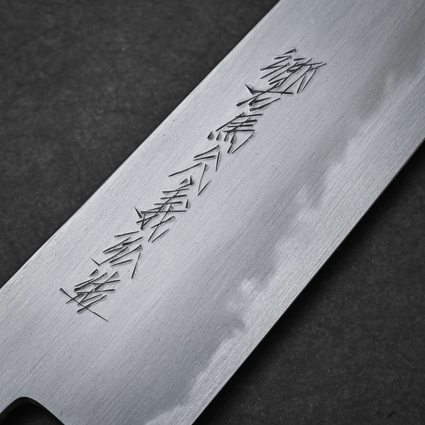A close-up of the Yoshikazu Tanaka shirogami#1 kiritsuke gyuto 225mm knife blade with engraved Japanese characters, crafted in Sakai, Japan, set against a dark textured background. The polished finish shows subtle reflections.