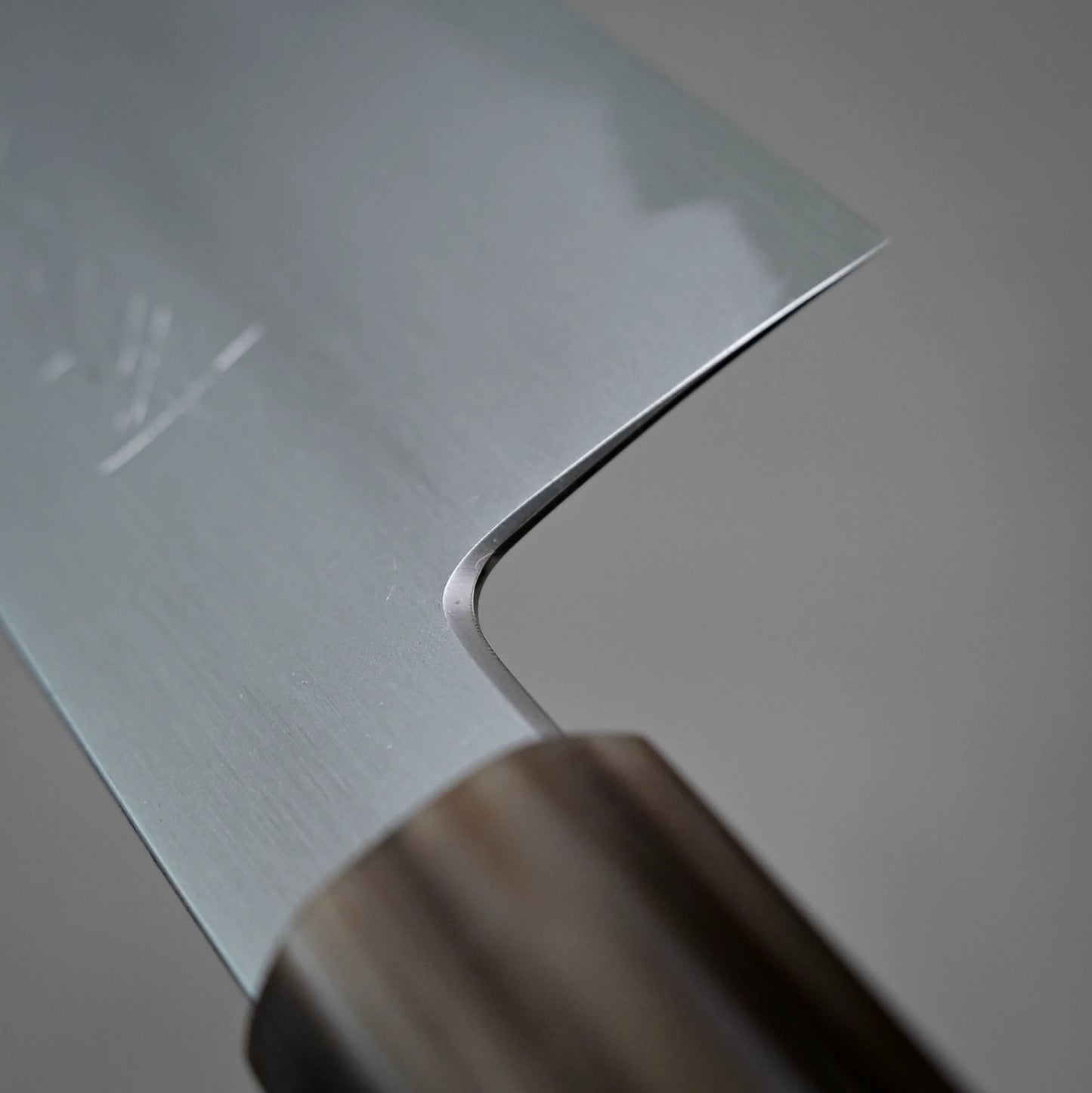 Close-up of the Yoshikazu Tanaka shirogami#1 kiritsuke gyuto 225mm kitchen knife, highlighting its smooth metal blade and wooden handle against a blurred gray background. Crafted in Sakai, Japan by Yoshikazu Tanaka.