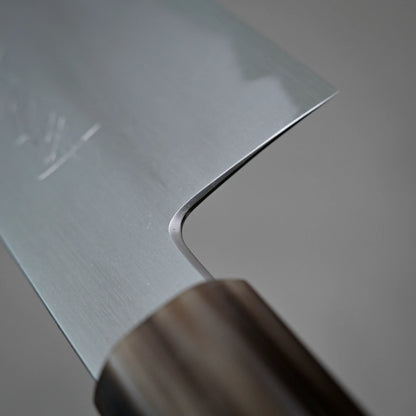 Close-up of the Yoshikazu Tanaka shirogami#1 kiritsuke gyuto 225mm kitchen knife, highlighting its smooth metal blade and wooden handle against a blurred gray background. Crafted in Sakai, Japan by Yoshikazu Tanaka.