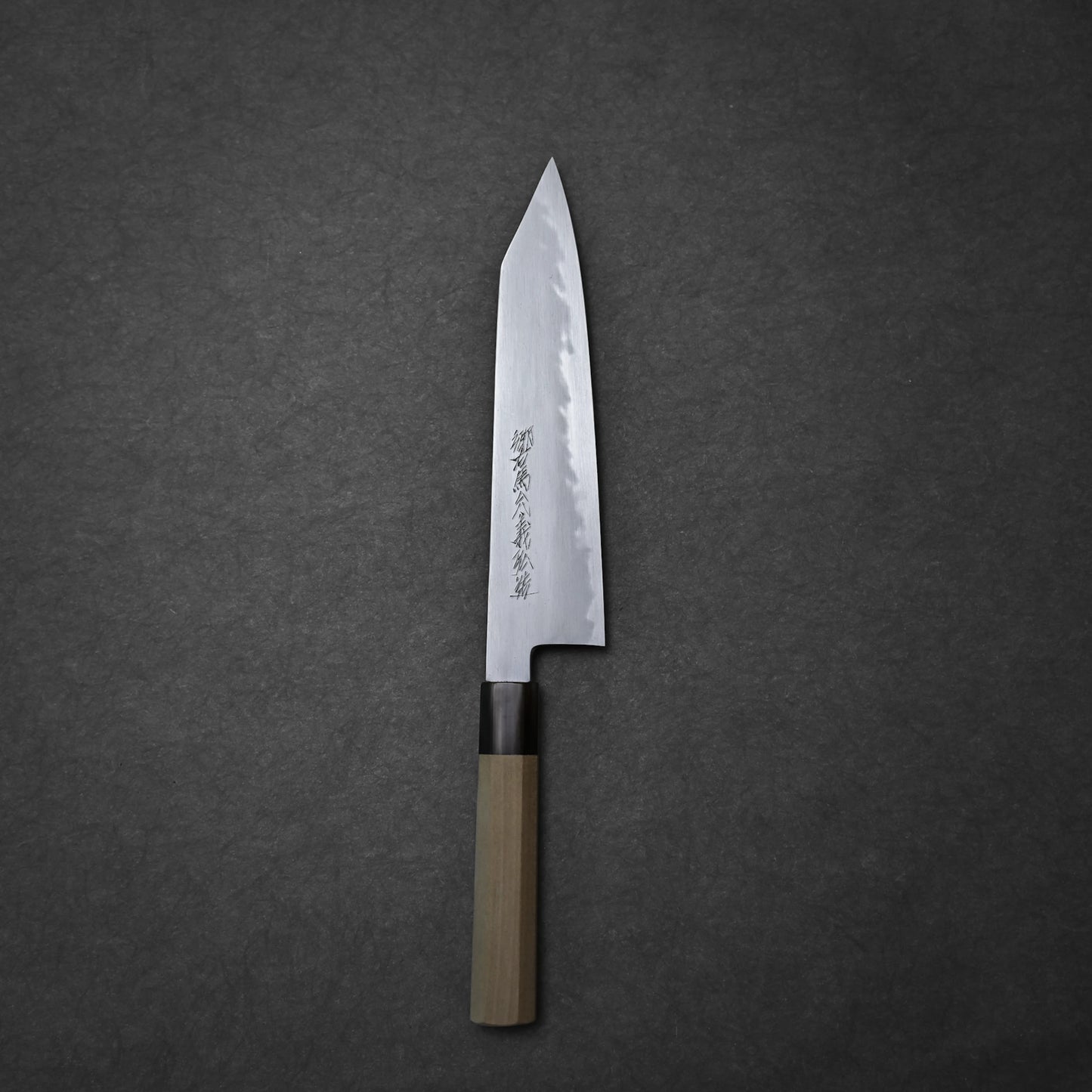The Yoshikazu Tanaka shirogami#1 kiritsuke gyuto 225mm, a Japanese chefs knife with a wooden handle and engraved blade, by Yoshikazu Tanaka of Sakai, Japan, rests on a dark textured surface.