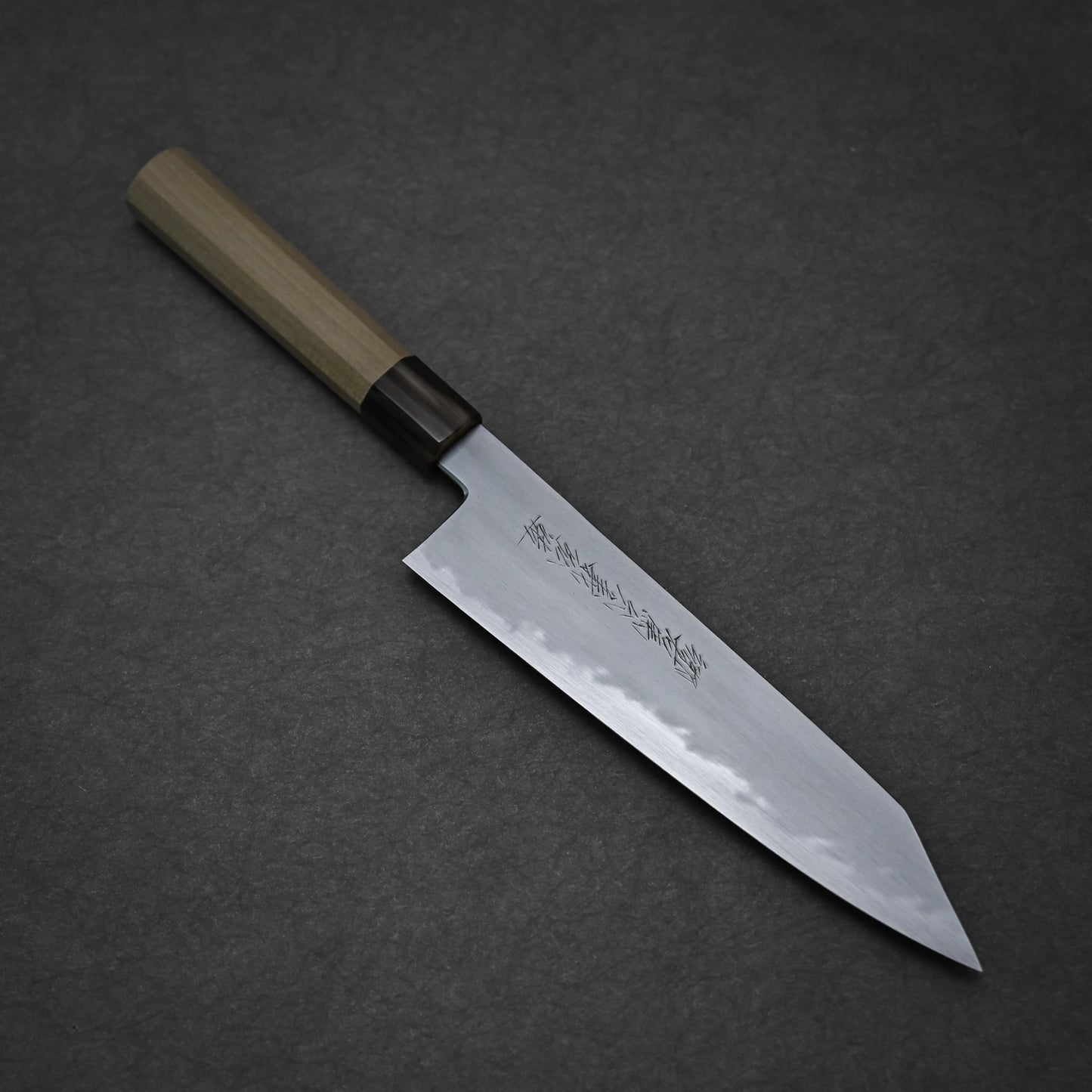 The Yoshikazu Tanaka shirogami#1 kiritsuke gyuto 225mm is a Japanese-style kitchen knife with a wooden handle and pointed steel blade engraved with Japanese characters, crafted by Yoshikazu Tanaka and shown on a dark textured surface.