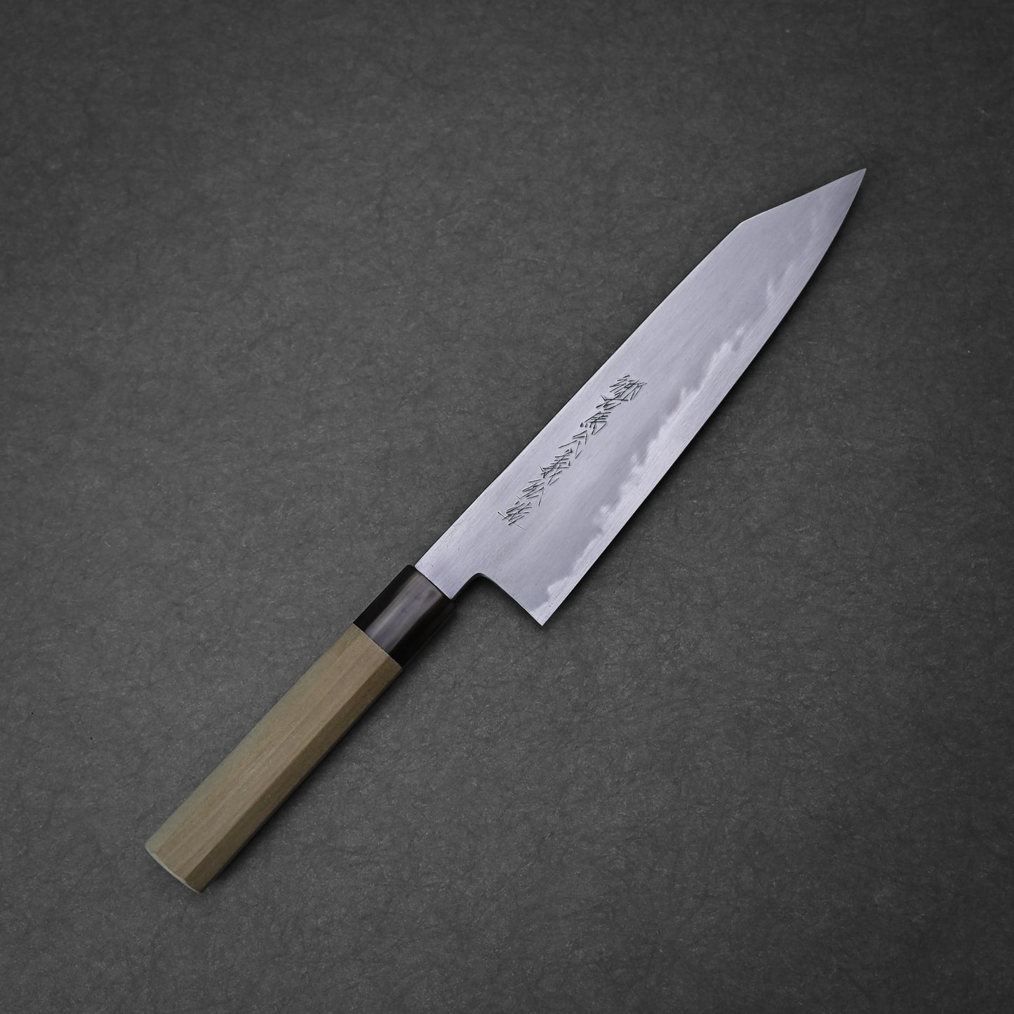 The Yoshikazu Tanaka shirogami#1 kiritsuke gyuto 225mm, featuring a wooden handle and an engraved pointed blade, rests on a dark gray textured surface.