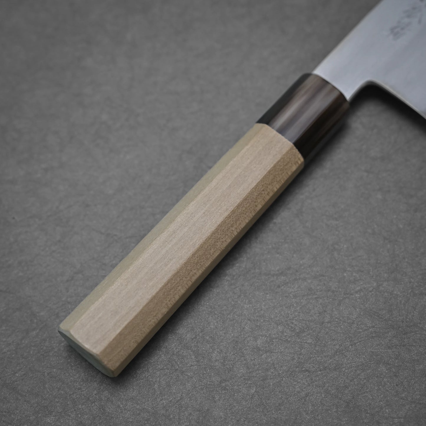 Close-up of the handle and blade of a Yoshikazu Tanaka shirogami#1 kiritsuke gyuto 225mm kitchen knife. The light wood handle has a dark section near the blade, crafted in Sakai, Japan, shown against a plain gray background.