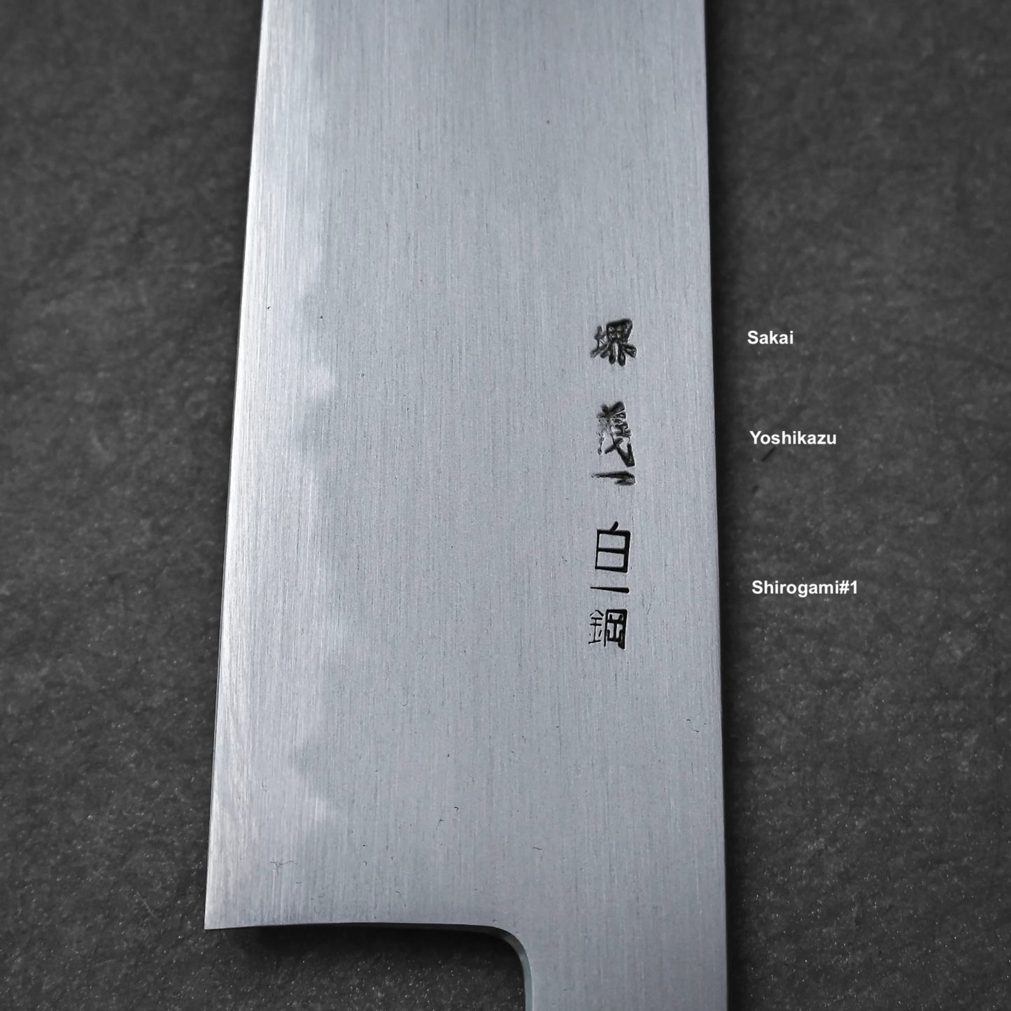 A close-up of the Yoshikazu Tanaka shirogami#1 kiritsuke gyuto 225mm blade shows engraved kanji. To the right, English labels display: Yoshikazu Tanaka and Shirogami #1. The knife rests on a dark textured surface.