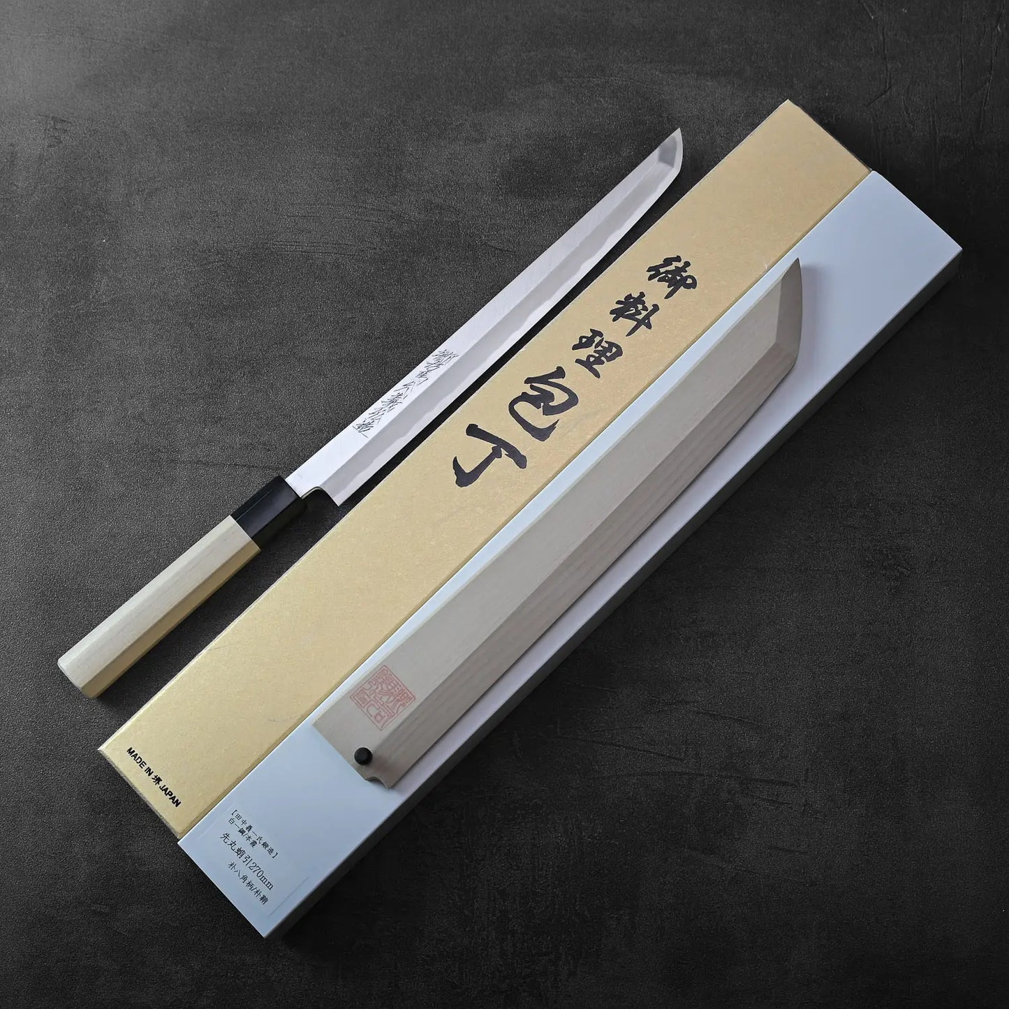 A sakimaru sujihiki with saya rests beside its box, showcasing refined lines and a harmonious silhouette.