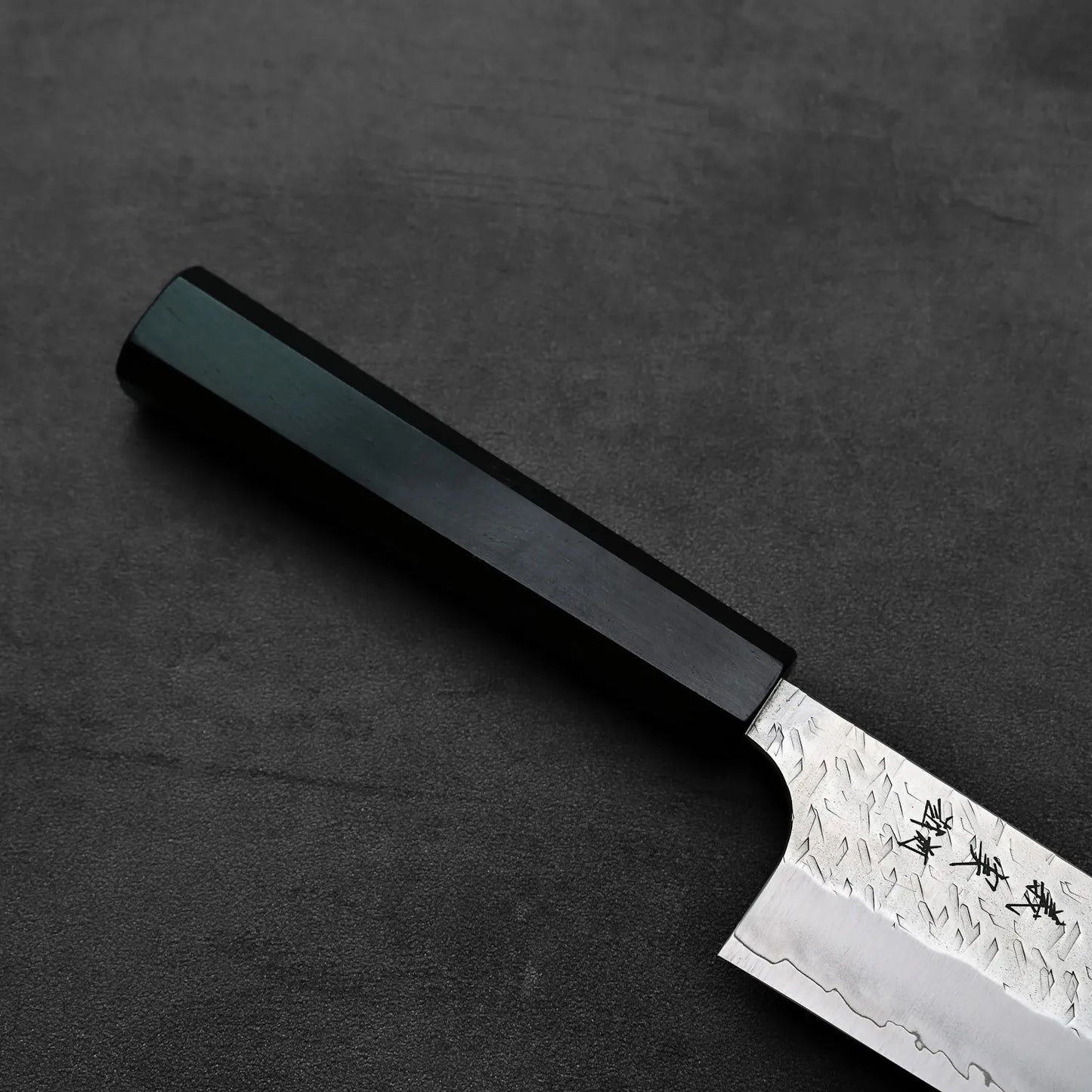 Kiritsuke gyuto featuring a dark handle and a textured blade viewed in close-up.