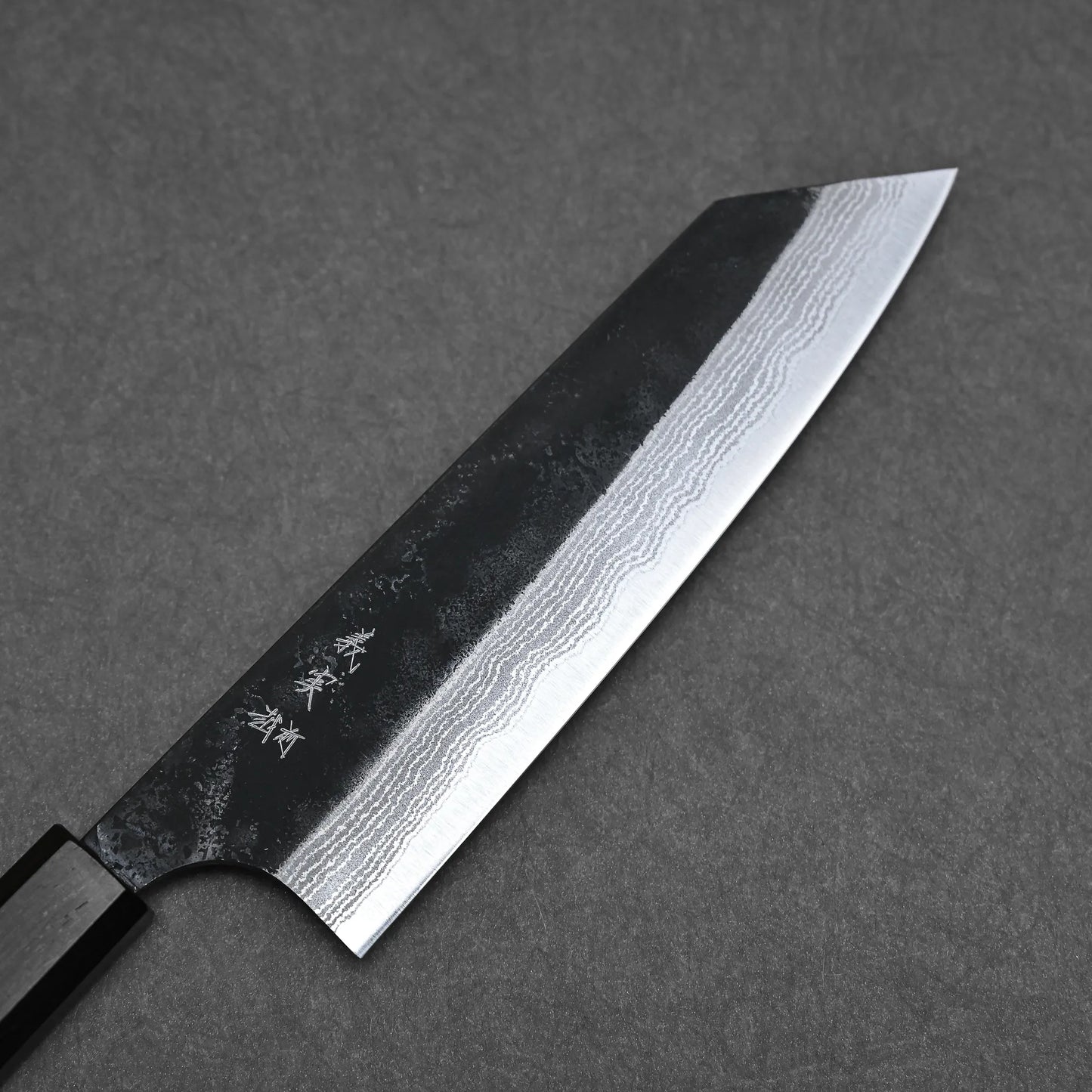 A Yoshimi Kato kurouchi damascus AS bunka 170mm knife with a dark handle and patterned blade, showing wavy layers and engraved characters, rests on a dark gray textured surface.