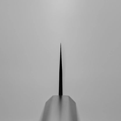 Close-up, symmetrical view of the Yoshimi Kato kurouchi damascus AS kiritsuke gyuto 210mm blade tip, centered and in focus against a plain light grey background; the knife’s edge is sharp while the rest appears blurred.