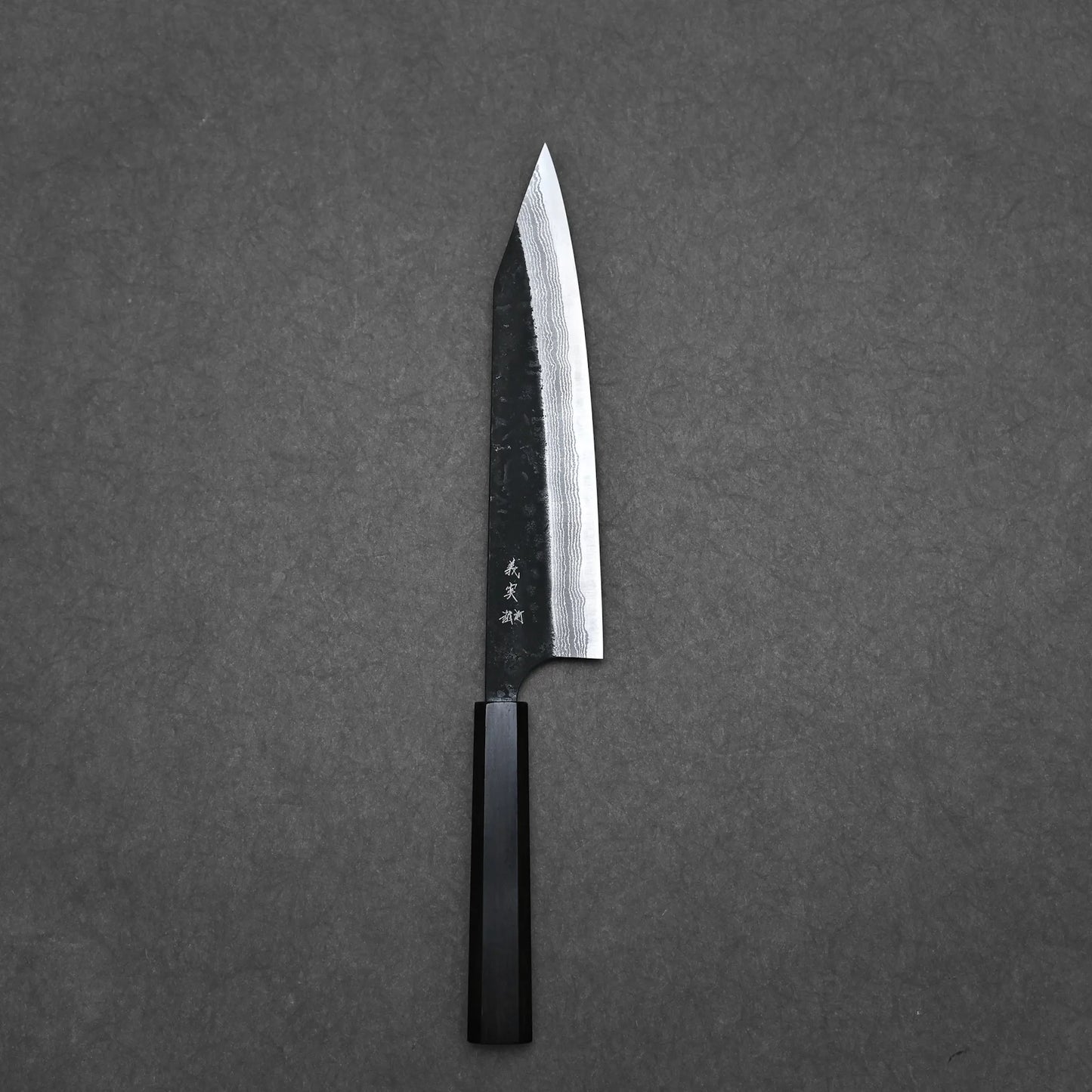 A Yoshimi Kato kurouchi damascus AS kiritsuke gyuto 210mm kitchen knife with a dark octagonal handle and sharp pointed blade rests on a plain dark gray surface.