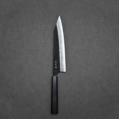 A Yoshimi Kato kurouchi damascus AS kiritsuke gyuto 210mm kitchen knife with a dark octagonal handle and sharp pointed blade rests on a plain dark gray surface.