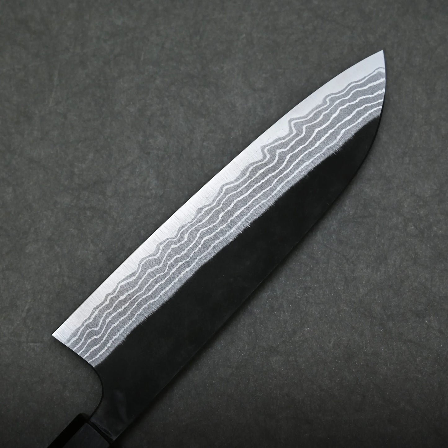 A close-up of a damascus santoku knife with a patterned Damascus steel blade, showing distinct wavy lines, set against a dark, textured background.