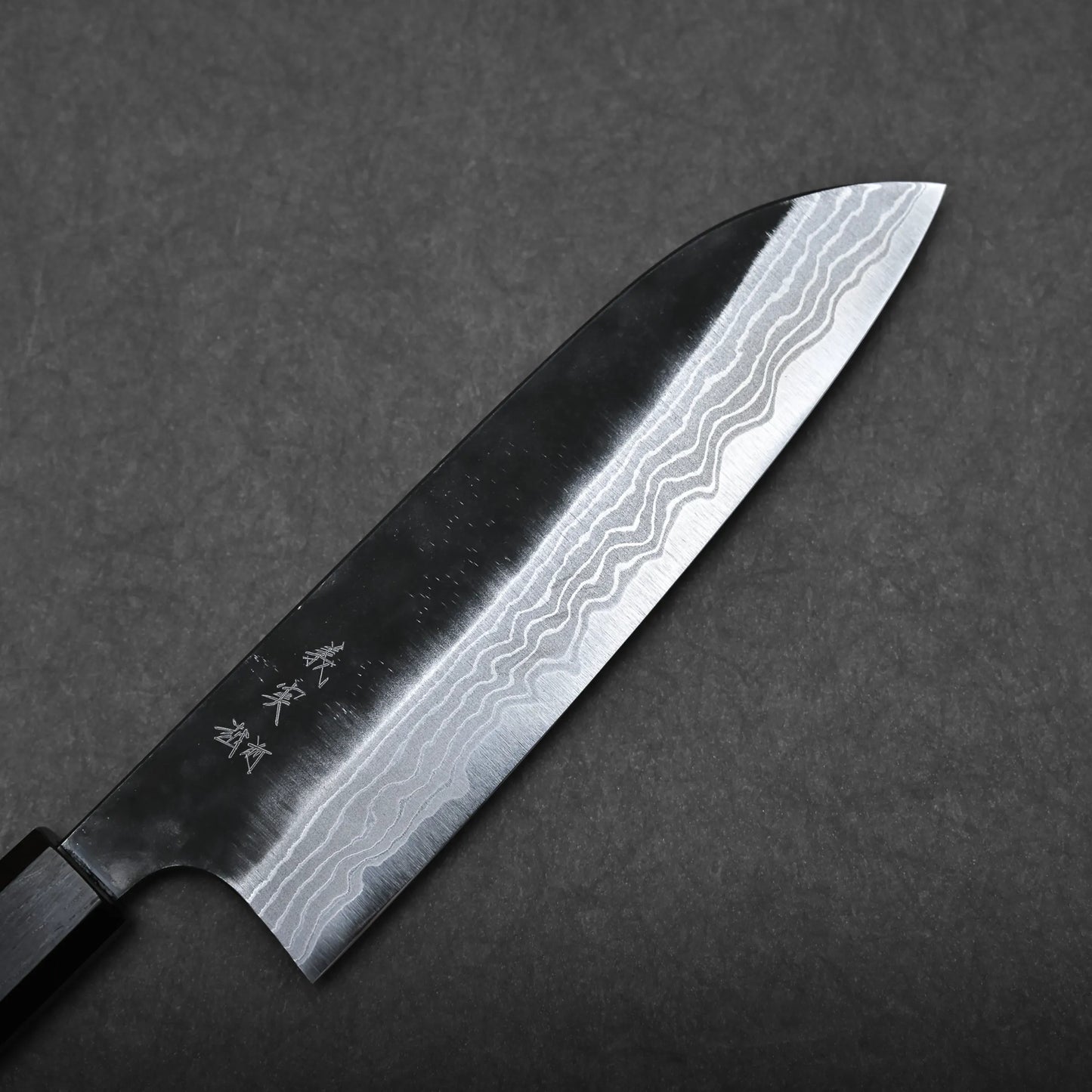 A Kato santoku knife with a dark handle and a blade featuring visible wavy Damascus steel patterns is positioned on a textured dark gray surface.