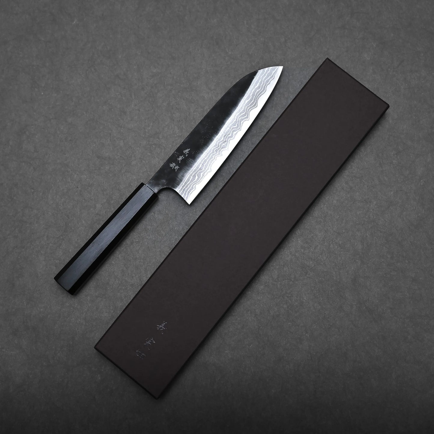 A Kato santoku knife with a dark handle and a patterned steel blade rests beside a closed, rectangular black box on a dark grey surface.