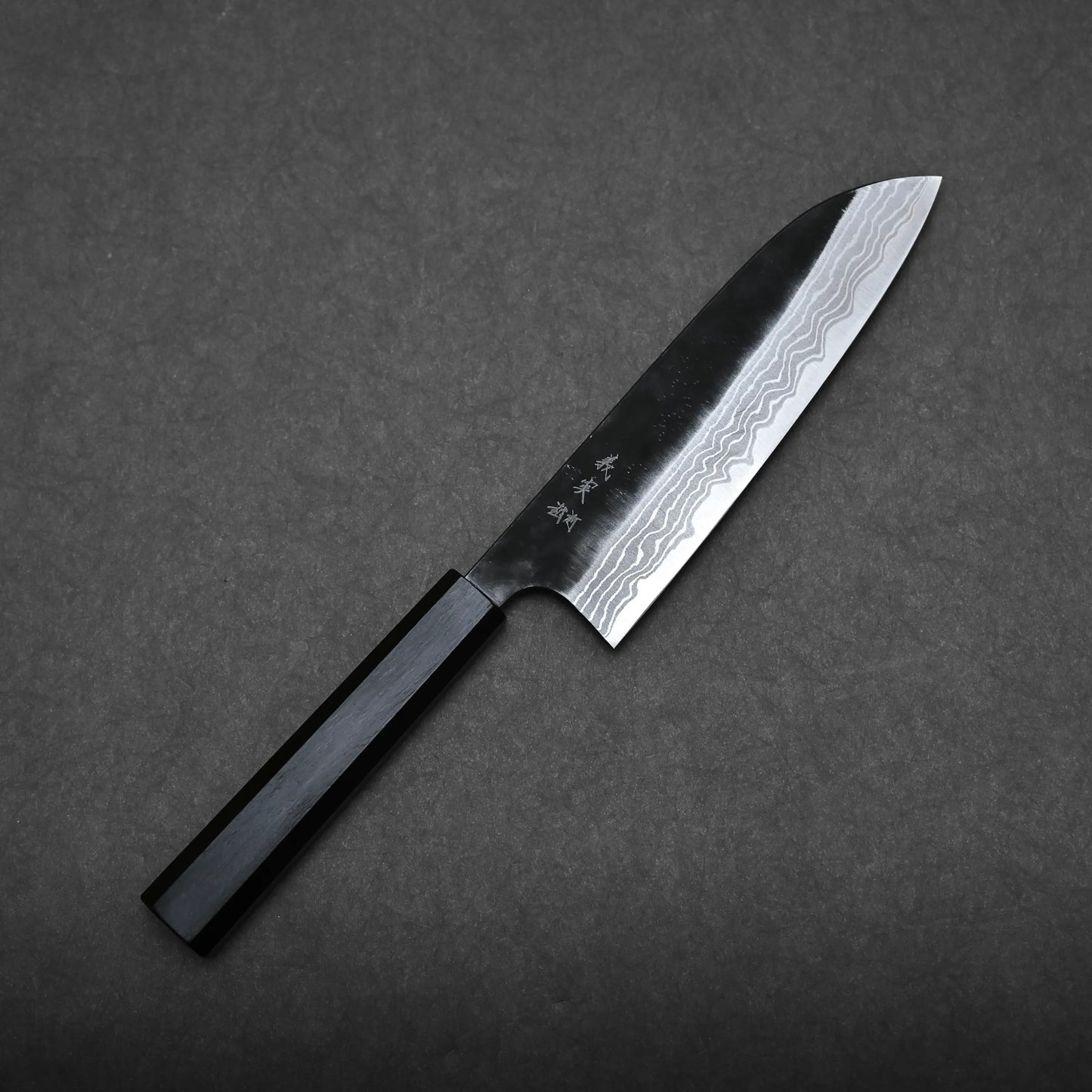 A Yoshimi Kato kurouchi damascus shirogami#2 santoku with a dark handle and a patterned, sharp blade rests on a plain dark gray background. The blade displays visible wavy lines and some engraved characters.