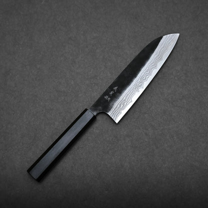 A Yoshimi Kato kurouchi damascus shirogami#2 santoku with a dark handle and a patterned, sharp blade rests on a plain dark gray background. The blade displays visible wavy lines and some engraved characters.