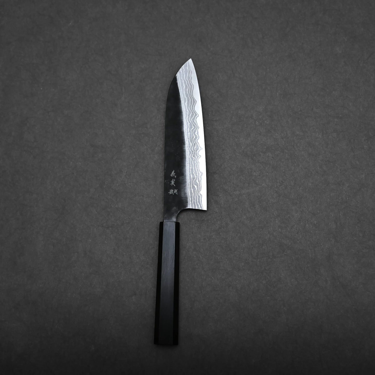 A Kato santoku knife with a patterned damascus blade and a dark handle lies on a flat, dark gray surface.