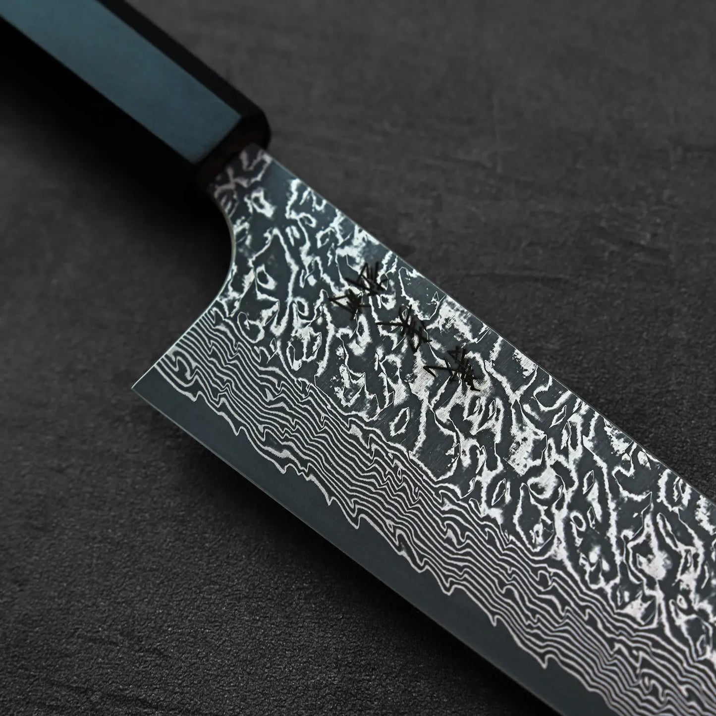 Three ants walk on a kiritsuke gyuto knife with a damascus blade and a black handle.