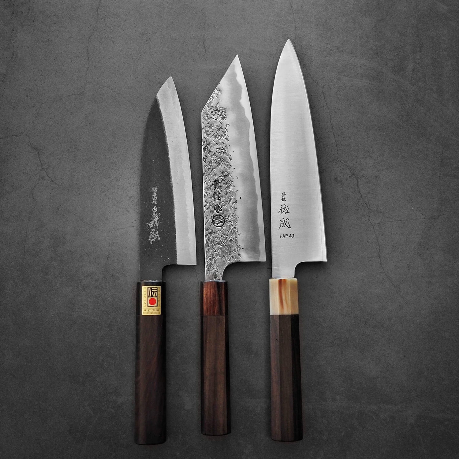 Top view of a trio of versatile Japanese knives including santoku, bunka, and gyuto are displayed. The Yoshihiro kurouchi santoku is positioned on the left, the Manaka tsuchime bunka in the middle, and the Sukenari Hap40 gyuto is showcased on the right.
