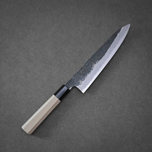 The Yoshikazu Tanaka Ittosai Kotetsu Gokujo kurouchi shirogami#1 gyuto 210mm, featuring a light wooden handle and dark blade, rests on a gray surface.