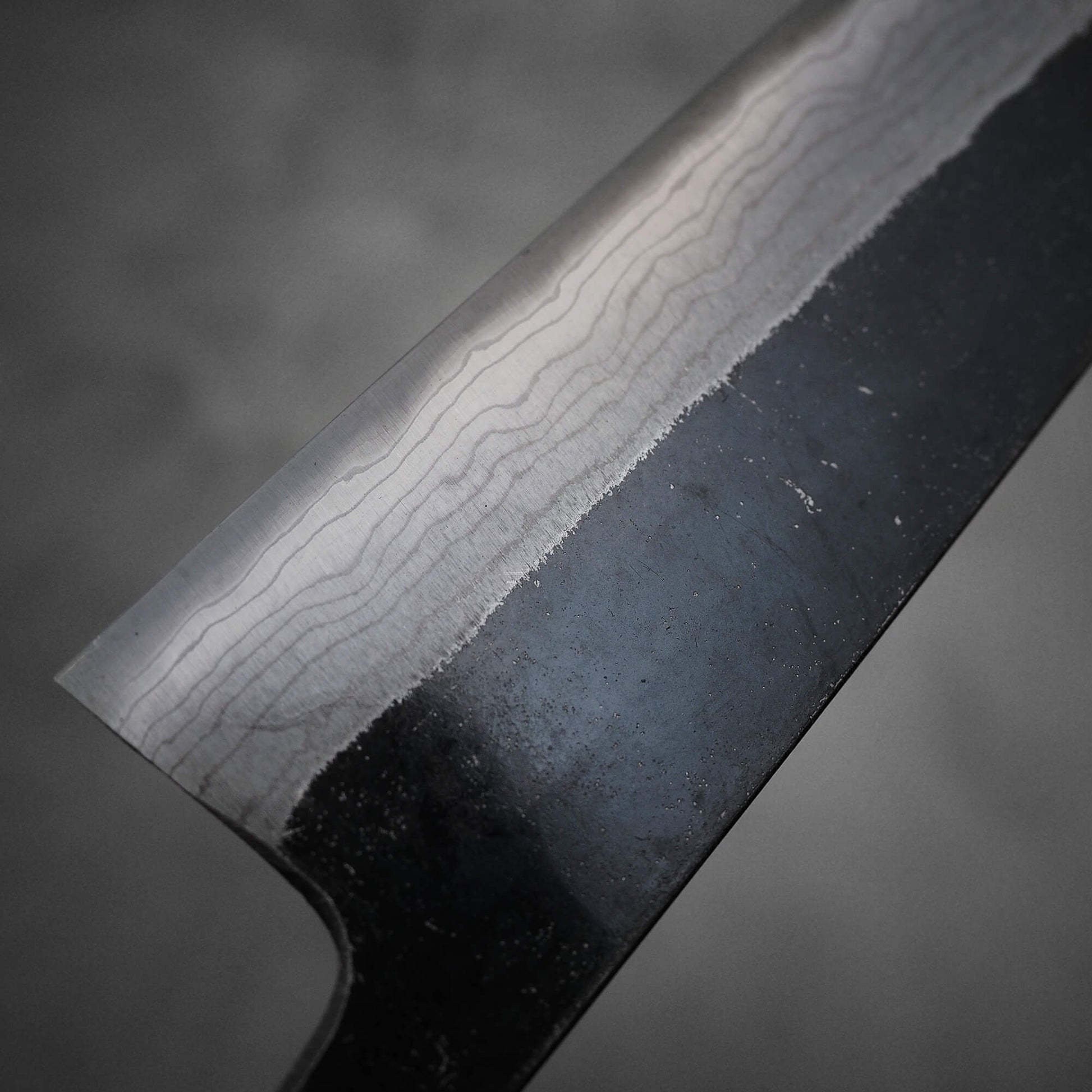 Close up view of Hatsukokoro Kumokage kurouchi damascus aogami#2 bunka knife. Image focuses on the left side of the blade at the heel