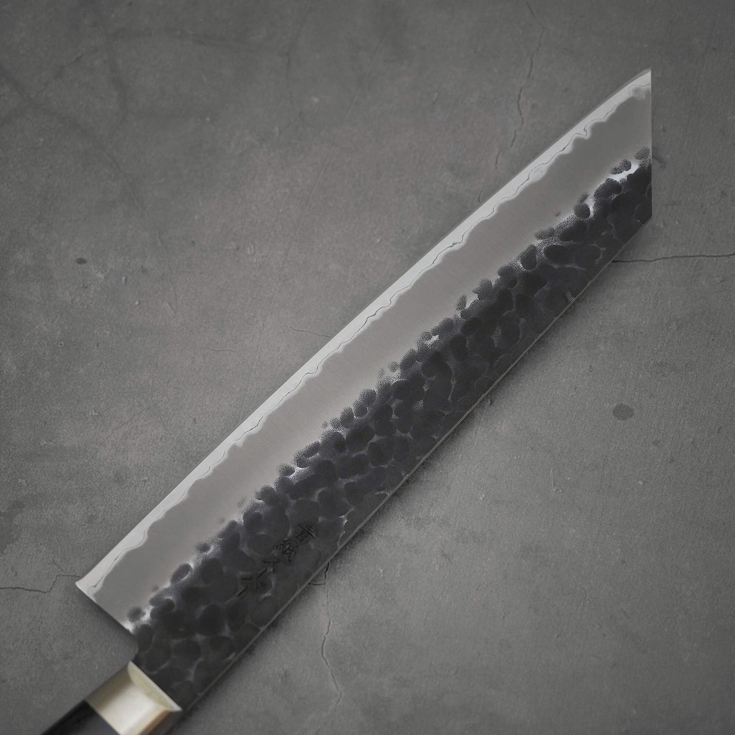 Top view of the blade of Ittosai Kotetsu tsuchime kurouchi aogami super kiritsuke gyuto. Image focuses on the left side of the blade
