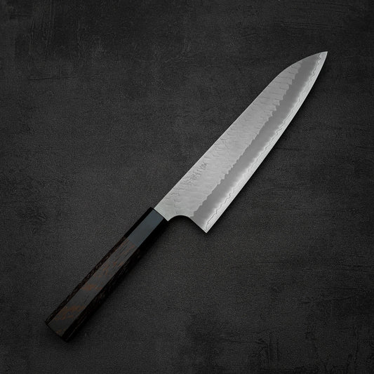 The Nigara Hamono Nigara tsuchime SG2 gyuto 210mm, featuring a dark wooden handle and a patterned SG2 steel blade, rests on a dark, textured surface.
