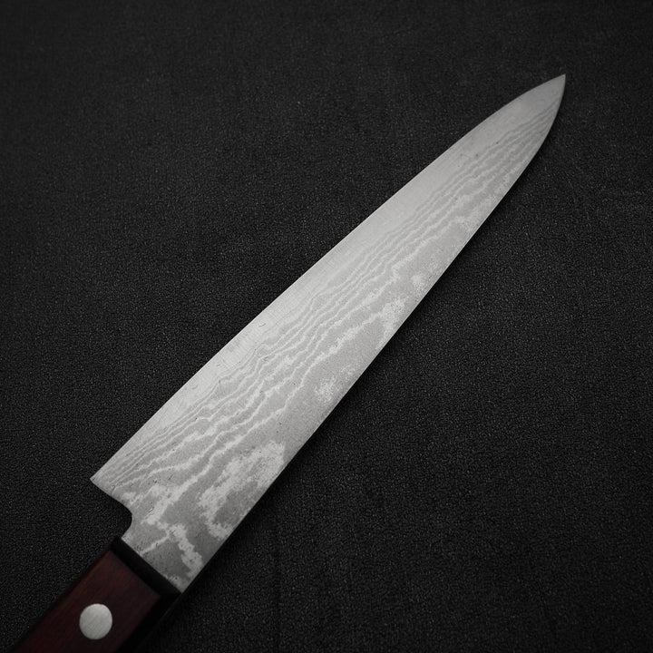 A close-up of the Shigeki Tanaka VG10 damascus 150mm petty knife, featuring a dark wooden handle and patterned stainless steel blade, positioned diagonally on a black textured surface.