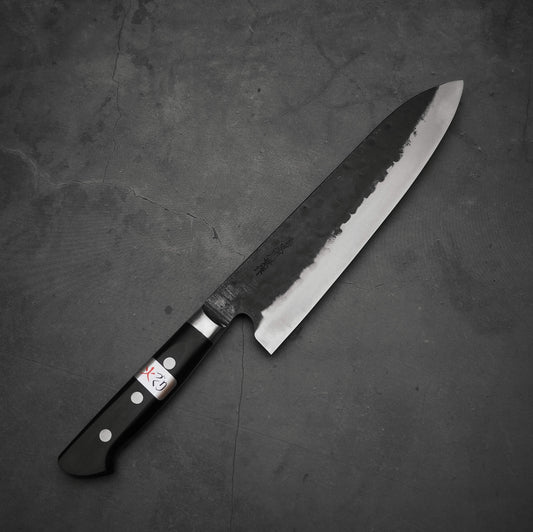A Teruyasu Fujiwara Denka gyuto 210mm knife with a black handle and partially polished blade, bearing visible markings and a small X23 sticker on the handle, rests on a dark textured surface.