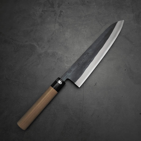 The Toyama kurouchi aogami#2 240mm gyuto chef’s knife by Toyama, featuring a wooden handle and a polished blade, rests on a gray textured surface.