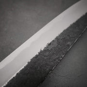 Close-up of a Yoshikazu Tanaka kurouchi AS gyuto 210mm, showcasing its polished Aogami Super edge and textured dark spine, resting on a matte surface.