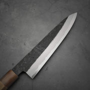 A Yoshikazu Tanaka kurouchi AS gyuto 210mm with a dark, textured blade and wooden handle rests on a gray, cracked concrete surface.