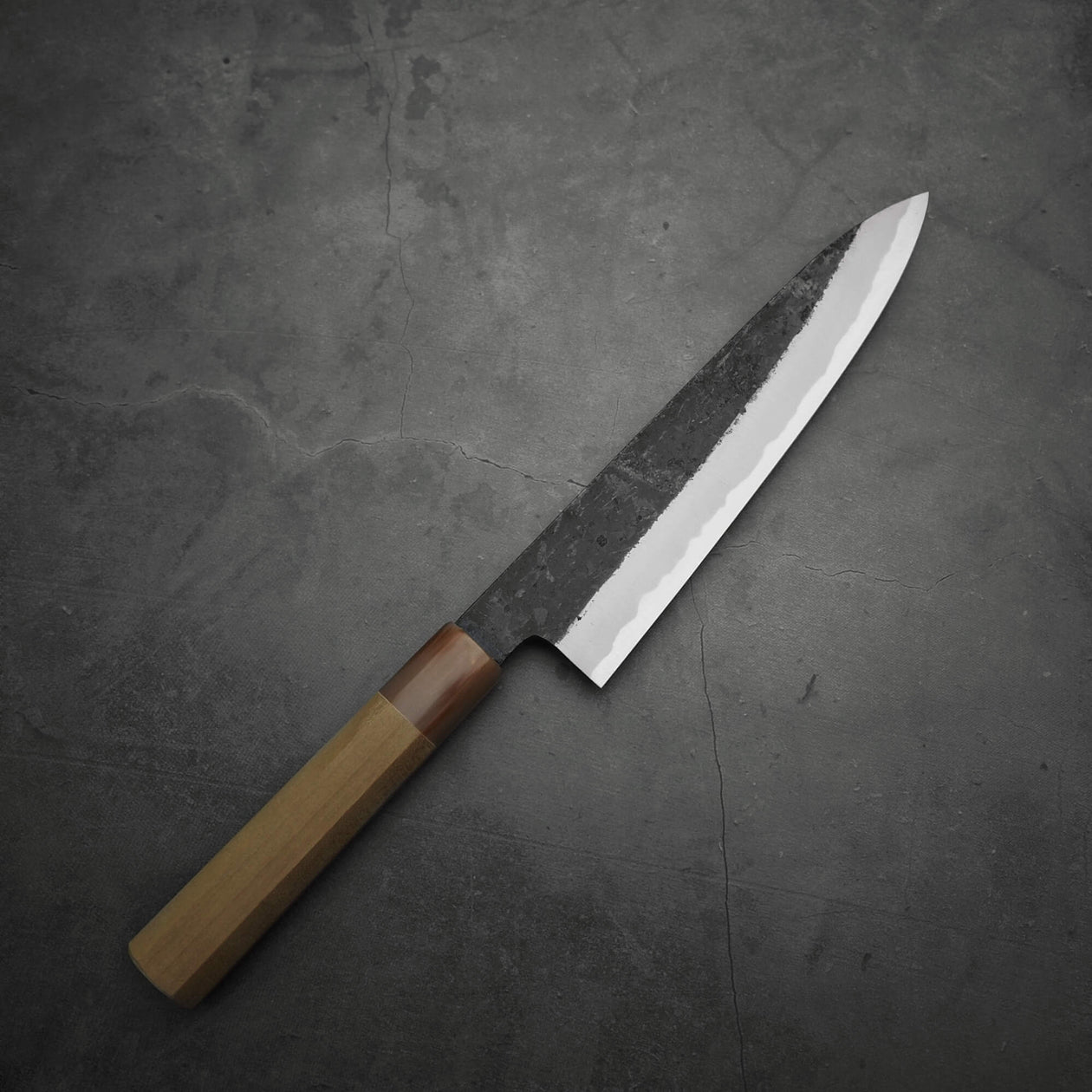 A Yoshikazu Tanaka kurouchi AS gyuto 210mm with a wooden handle sits on a dark, textured stone surface.