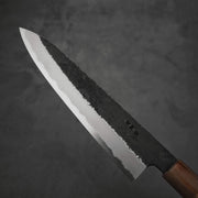 A close-up of the Yoshikazu Tanaka kurouchi AS gyuto 210mm chef’s knife, featuring a hammered Aogami Super blade and wooden handle by Yoshikazu Tanaka, shown against a dark, textured background.