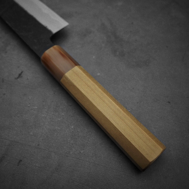 Close-up of a Yoshikazu Tanaka kurouchi AS gyuto 210mm handle in light wood with a darker section near the blade, set on a dark background. The visible blade edge hints at its Aogami Super steel construction.