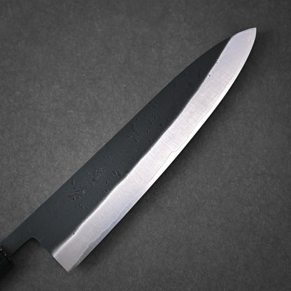 The Ohishi kurouchi SLD gyuto 210mm by Ohishi, featuring a dark handle and polished blade with water droplets, rests on a textured surface.