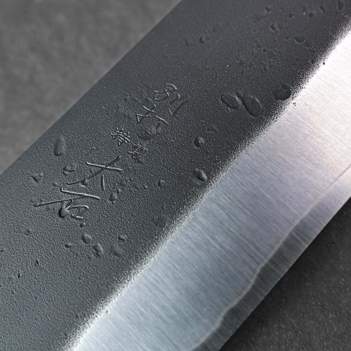 Close-up of the Ohishi kurouchi SLD gyuto 210mm blade by Ohishi, showing water droplets and engraved Japanese characters on the dark metal, and emphasizing the polished edge contrasted with its matte texture.