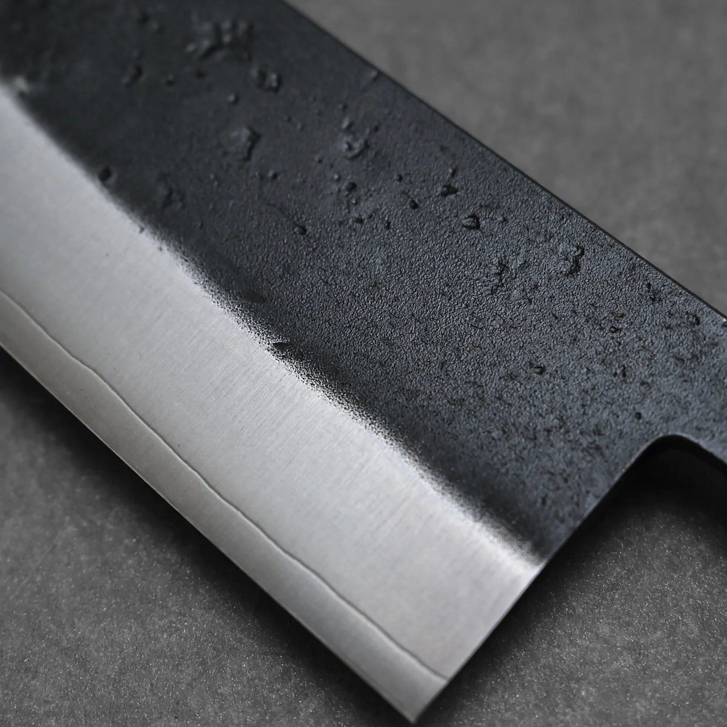 Close-up of the Ohishi kurouchi SLD gyuto 210mm by Ohishi, highlighting its sharp edge and dark, textured blade with subtle irregular spots against a smooth, dark background.
