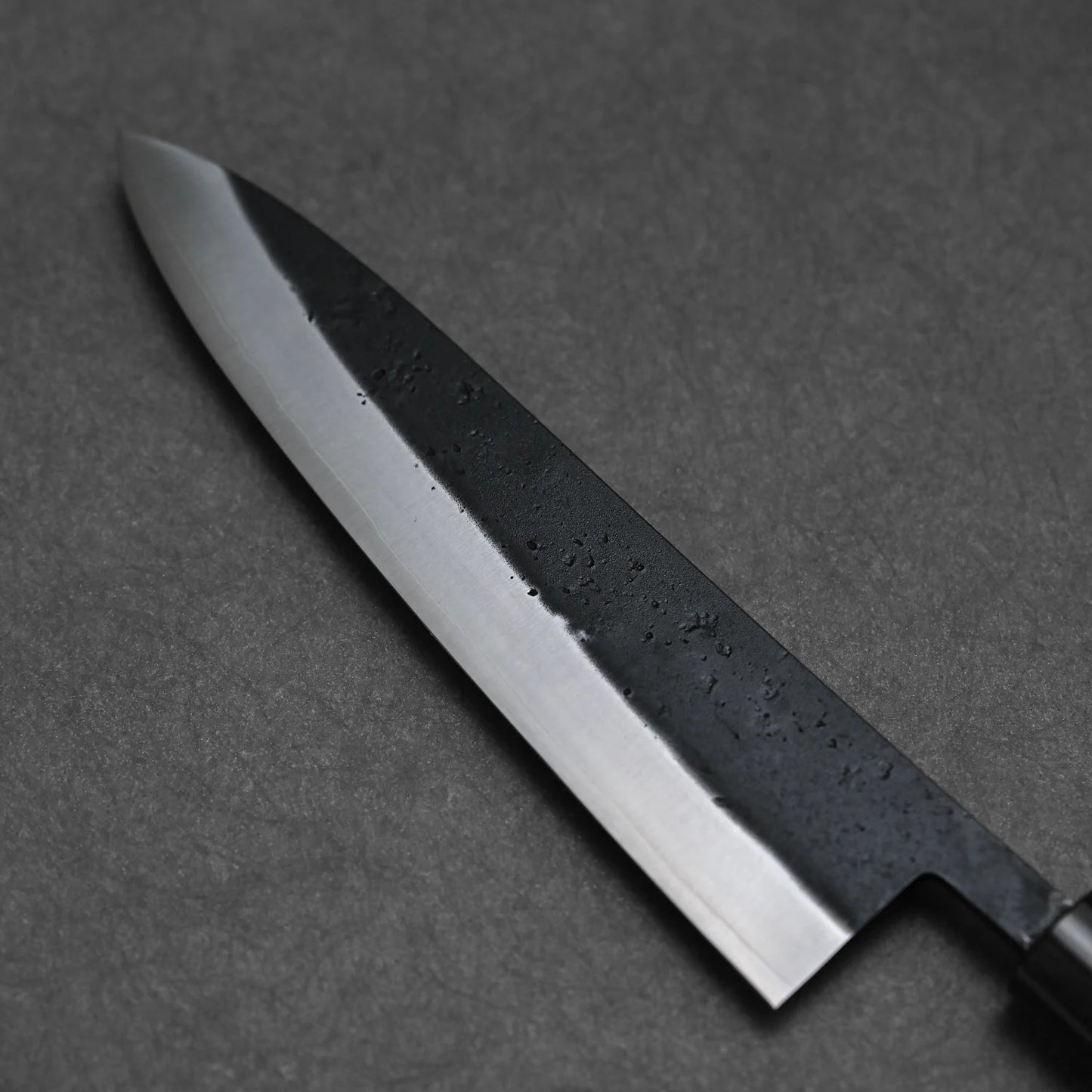 A close-up of the Ohishi kurouchi SLD gyuto 210mm Japanese chef knife by Ohishi, featuring a polished blade with a dark, textured section near the spine, displayed on a dark gray surface.
