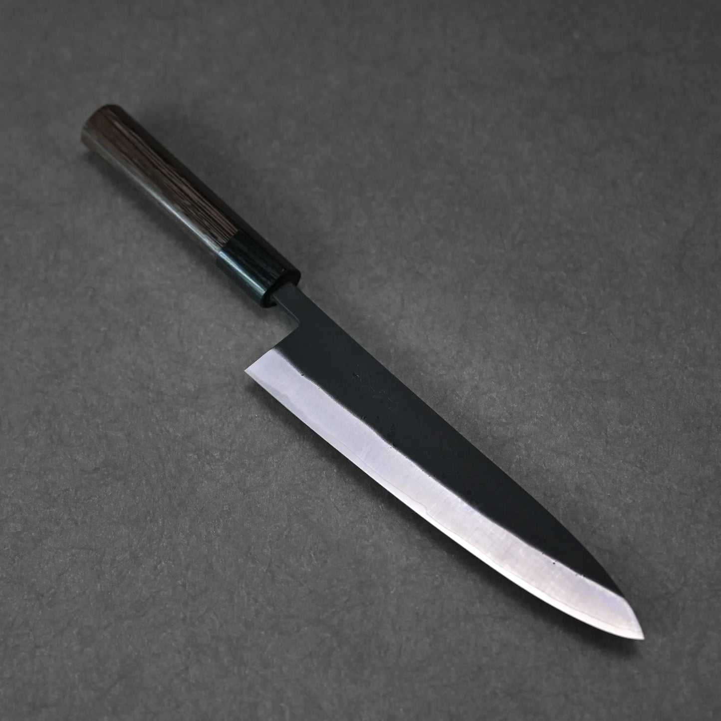 The Ohishi kurouchi SLD gyuto 210mm chef knife by Ohishi, featuring a dark wooden handle and sharp, shiny blade, rests on a dark, textured surface.