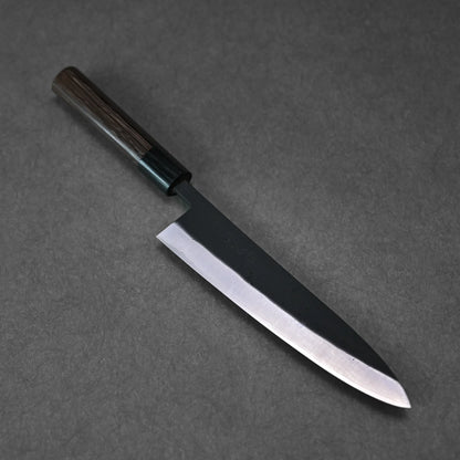 The Ohishi kurouchi SLD gyuto 210mm chef knife by Ohishi, featuring a dark wooden handle and sharp, shiny blade, rests on a dark, textured surface.