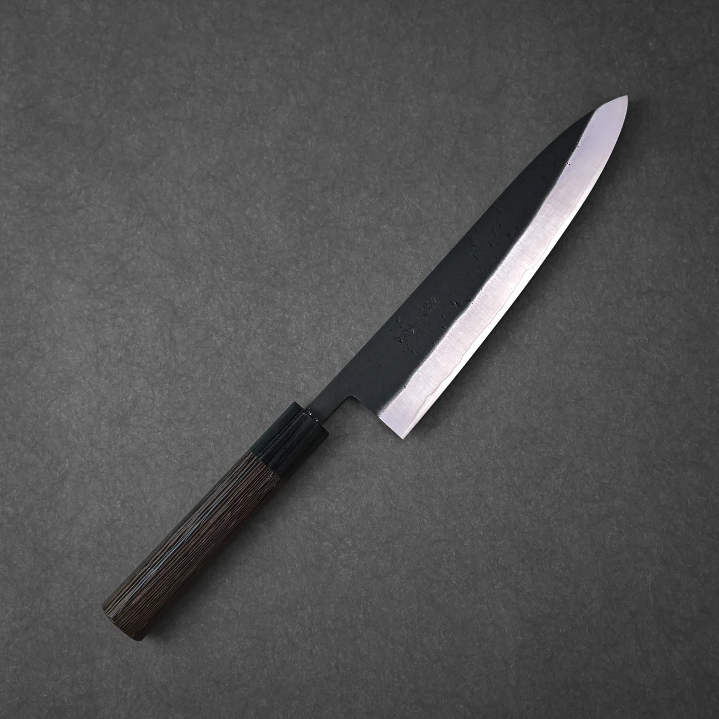 The Ohishi kurouchi SLD gyuto 210mm by Ohishi features a dark wooden handle and a black blade with a polished edge, shown on a dark gray textured surface.