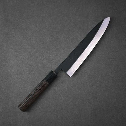 The Ohishi kurouchi SLD gyuto 210mm by Ohishi features a dark wooden handle and a black blade with a polished edge, shown on a dark gray textured surface.
