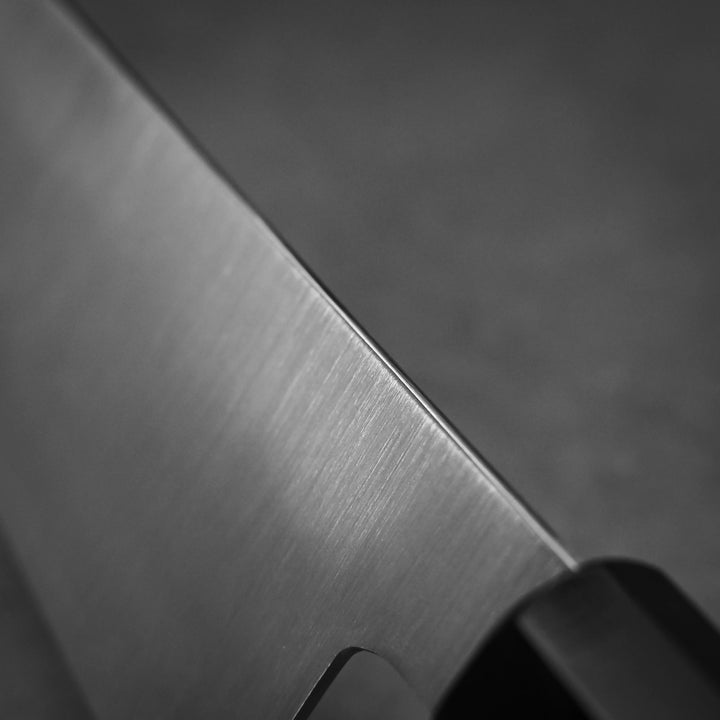 Close-up of the sharp edge of a Shigeki Tanaka ginsan gyuto 240mm Japanese kitchen knife with a Ginsan stainless steel blade and black handle, set against a blurred gray background. Brand: Shigeki Tanaka.