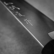 Black and white close-up of the Shigeki Tanaka ginsan gyuto 240mm, featuring Japanese characters engraved on its Ginsan stainless steel blade. The blurred background highlights the fine edge and precise details.