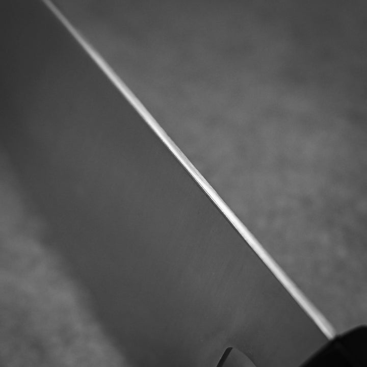 A close-up black and white photo captures the sharp edge of the Shigeki Tanaka ginsan gyuto 240mm kitchen knife, showing part of its handle on the right against a blurred background.
