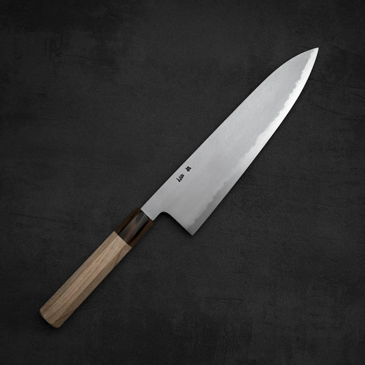 The Yoshikazu Tanaka AS tall gyuto 240mm knife, featuring a wooden handle and a sharp stainless steel blade by Yoshikazu Tanaka, is placed diagonally on a dark, textured surface.