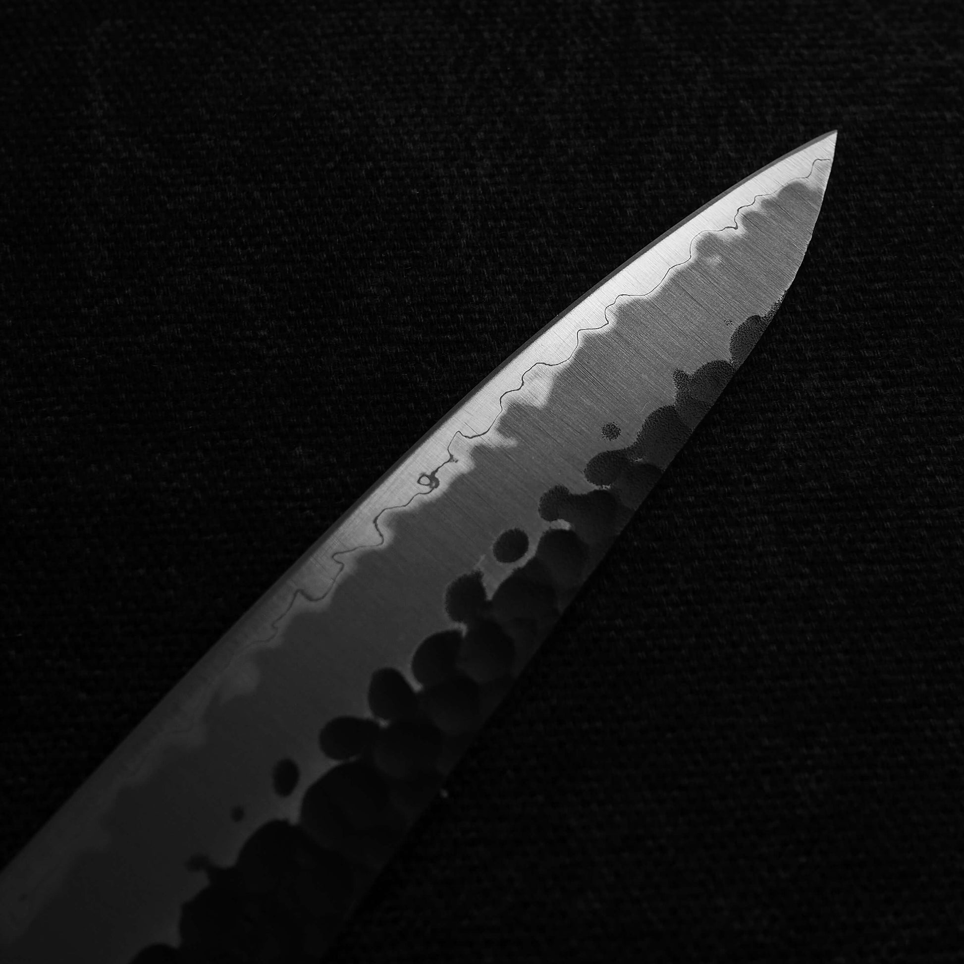 Close up view of Ittosai Kotetsu tsuchime kurouchi aogami super petty knife. Image focuses on the tip area of the left side of the knife