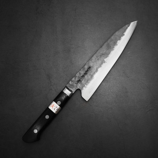 A Teruyasu Fujiwara Denka gyuto 180mm with a black, three-rivet handle rests on a dark textured surface. Its Aogami Super steel blade features a hammered finish and Japanese characters near the handle.