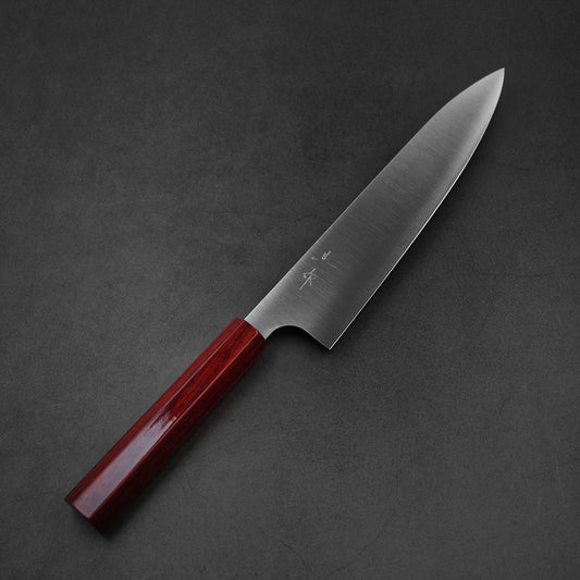 A Kei Kobayashi SG2 gyuto 210mm Japanese kitchen knife with a polished dark red wooden handle rests on a black textured surface, its blade facing right.